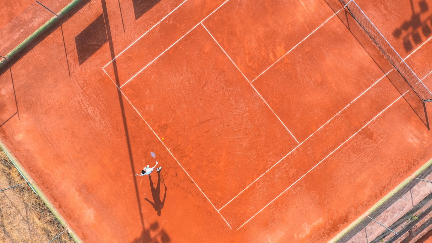 Jugador de tenis golpeando la pelota en una cancha de arcilla roja con sombras largas.