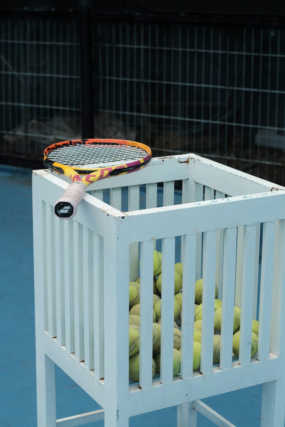 Raqueta de tenis apoyada en un carrito lleno de pelotas de tenis.